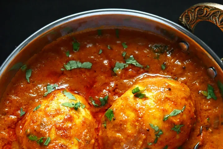 Close-up of spiced eggs in rich tomato curry garnished with herbs.